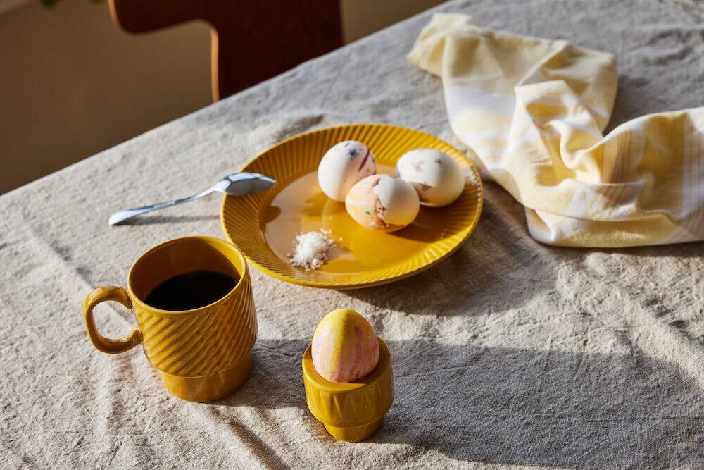 Auf einer beigen Tischdecke stehen ein Kaffeebecher der Sagaform GmbH Coffee & More, ein gelber Teller mit Ostereiern, ein Löffel, Salz und eine gestreifte Serviette. Im Vordergrund liegt ein dekoriertes Ei in einem gelben Eierbecher.