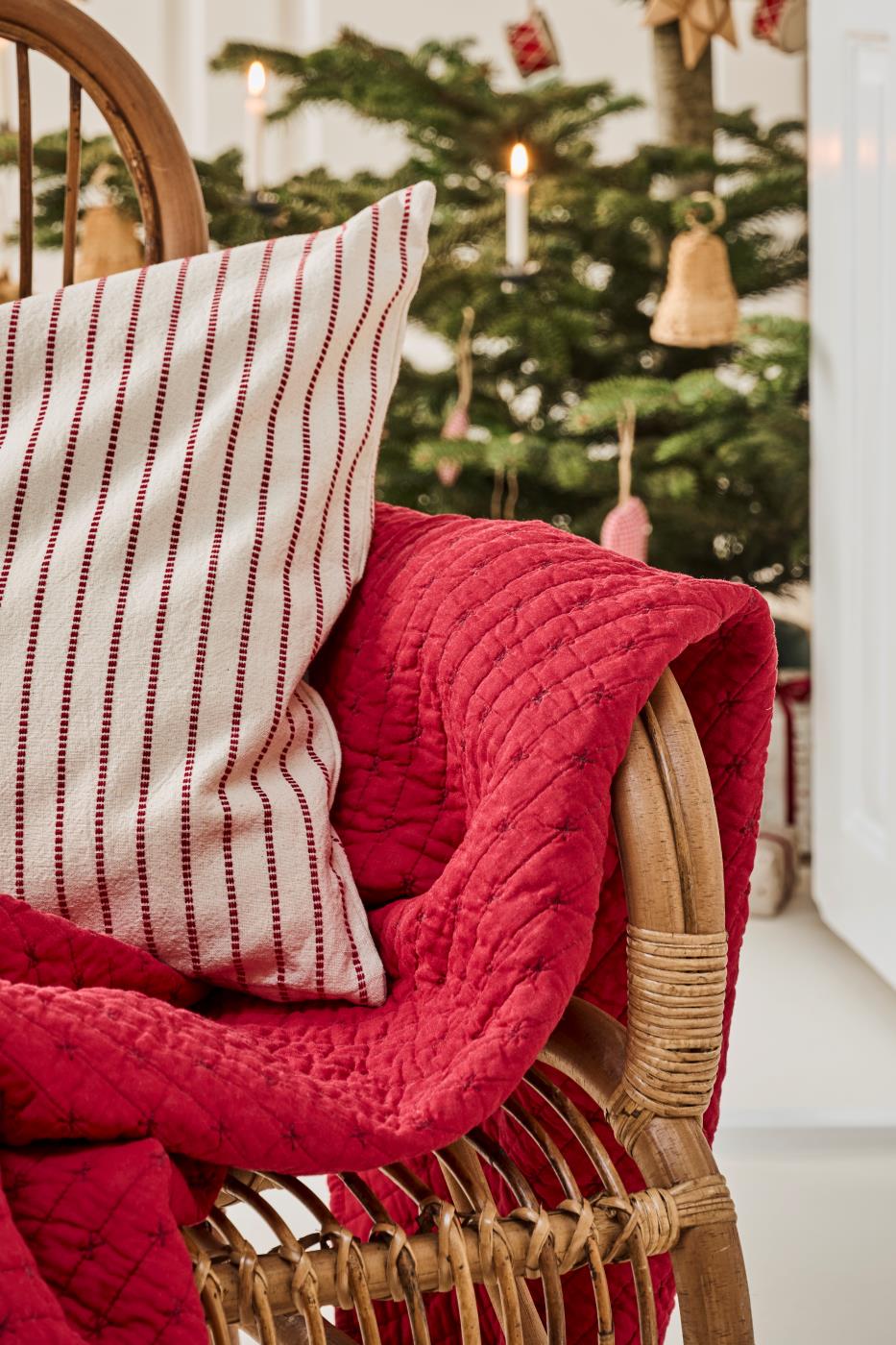 Ein Korbsessel mit einer IB Laursen Quilt Decke rot mit Kreuzstich und einem weißen Kissen mit roten Streifen steht vor einem mit Ornamenten und Lichtern geschmückten Weihnachtsbaum.