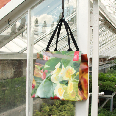 Die Upcycling KGA Tasche 'Blume' von KGA mit ihrem lebhaften Blumendesign hängt an einer Gewächshaustür, umgeben von üppigen Pflanzen und Glasplatten im Hintergrund.