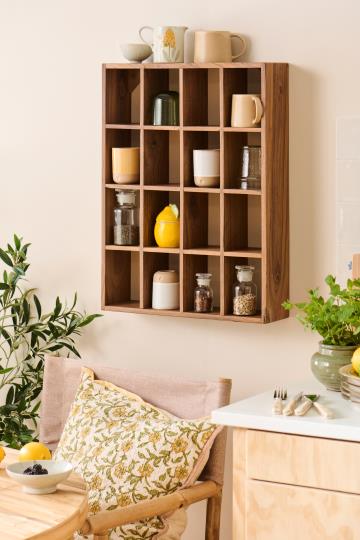 Ein hölzernes Wandregal mit Fächern bietet Platz für Tassen, die IB Laursen "Dose mit Deckel | Zitrone" und eine gelbe Teekanne über einem gemütlichen Stuhl mit Blumenkissen in einer luftigen Küche mit Grünpflanzen - perfekt für eine stilvolle Küchenaufbewahrung.