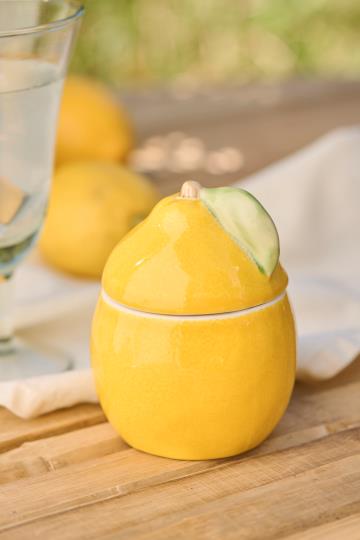 Die Dose mit Deckel | Zitrone von IB Laursen, perfekt für die Aufbewahrung in der Küche, steht auf einer hölzernen Oberfläche neben einem Glas Wasser, mit verschwommenen Zitronen und einem weißen Tuch im Hintergrund.