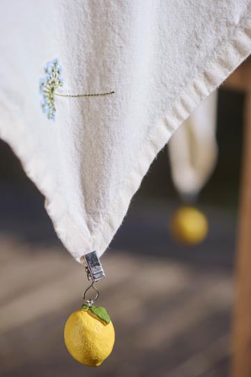 Ein weißes Tischtuch mit einer gestickten blauen Blume wird an der Ecke von einem IB Laursen Tischdeckenbeschwerer Schachtel Zitronengewicht (4 Stück Zitronen, 92294-00) umklammert, während im Hintergrund ein weiteres zitronenförmiges Gewicht hängt.