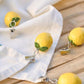 Vier IB Laursen Tischdeckenbeschwerer Schachtel in Form von Zitronen (4 Stück Zitronen) werden an der Ecke einer weißen Stoffserviette auf einer Holzfläche befestigt.