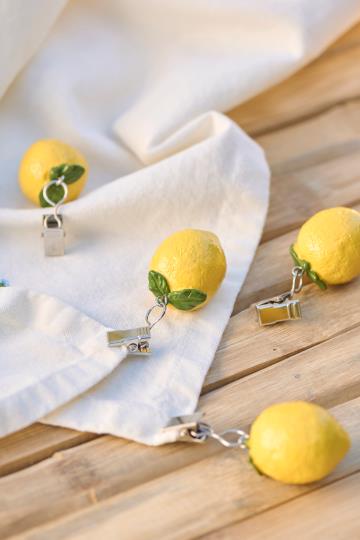 Vier IB Laursen Tischdeckenbeschwerer Schachtel in Form von Zitronen (4 Stück Zitronen) werden an der Ecke einer weißen Stoffserviette auf einer Holzfläche befestigt.