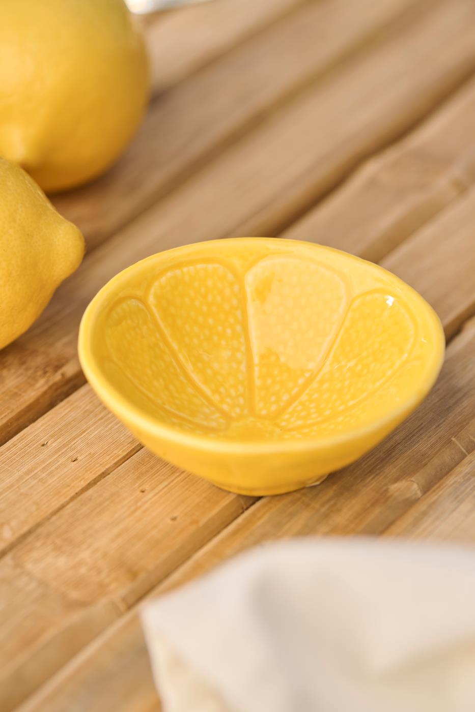 Die Schale | mini Zitrone von IB Laursen, eine gelbe Keramikschale mit dem Muster einer Zitronenscheibe, steht auf einer Holzfläche mit Zitronen in der Nähe und einem weißen Tuch im Vordergrund.