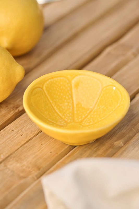 Die Schale | mini Zitrone von IB Laursen, eine gelbe Keramikschale mit dem Muster einer Zitronenscheibe, steht auf einer Holzfläche mit Zitronen in der Nähe und einem weißen Tuch im Vordergrund.