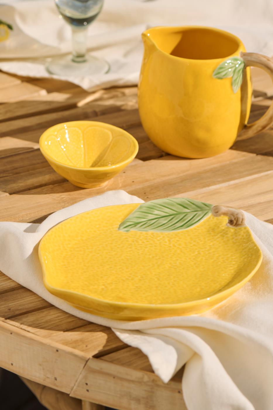 Ein gelber Keramikteller in Form einer Zitrone, die Schale | mini Zitrone von IB Laursen und ein gelber Krug mit Blattgriff stehen auf einem Holztisch, der mit einem weißen Tuch bedeckt ist.
