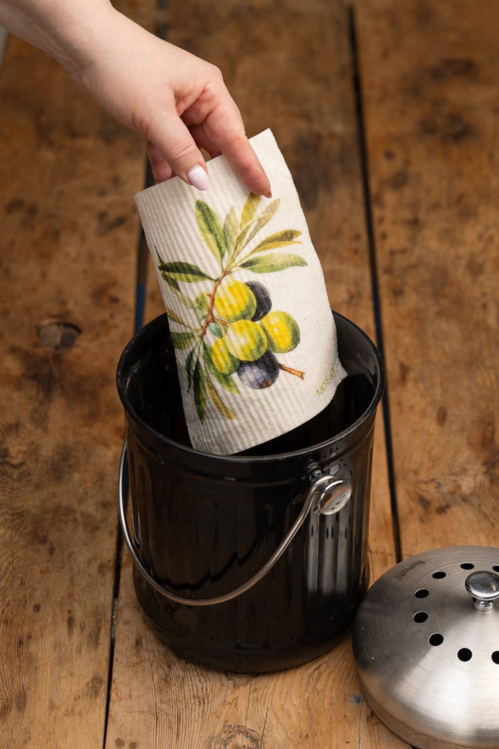Eine Hand legt das biologisch abbaubare Schwammtuch More Joy - Schwammtuch Olives mit Olivenzweigmotiv in eine schwarze Komposttonne auf einer Holzfläche. Der Metalldeckel liegt neben der Tonne.