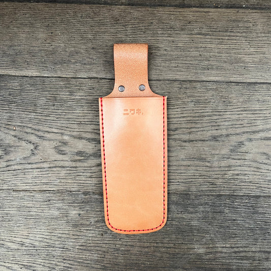 Ein hellbraunes Niwaki-Sägeholster von Niwaki mit roten Nähten und einem Schlaufengriff liegt flach auf Holz. Das Holster hat zwei Metallnieten an der Spitze und geprägte Branding auf der Vorderseite.