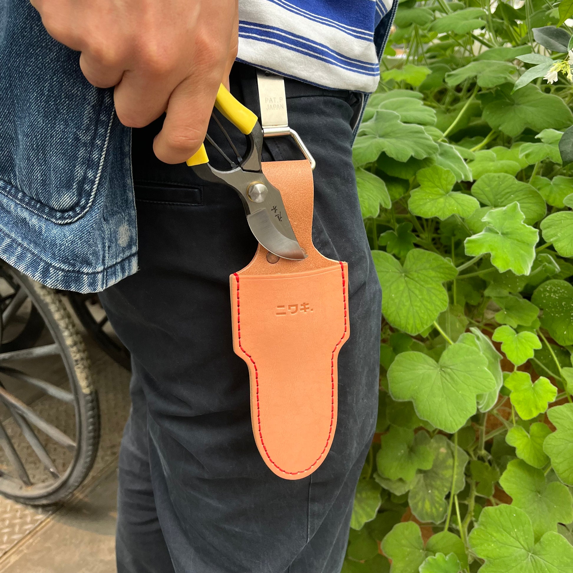 Eine Person in dunklen Hosen und einem blau gestreiften Hemd hält eine Gartenschere mit gelbem Griff über einem Niwaki-Gürtelclip von Niwaki, der an ihrem Gürtel befestigt ist, und steht neben grünen Blattpflanzen.