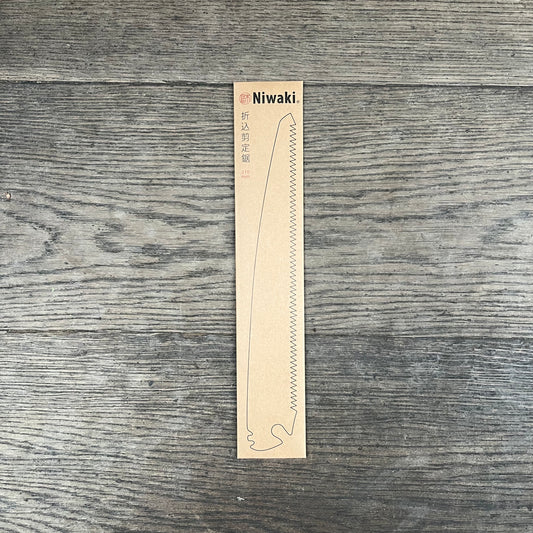 Eine Pappverpackung mit der Aufschrift Niwaki Ersatzklinge, mit der Marke Niwaki und einer schwarzen Umrisszeichnung sowie japanischem Text liegt auf einer Holzfläche.