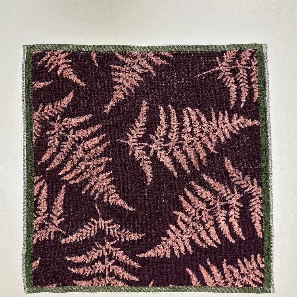 Das Küchenhandtuch "Verzauberter Wald" von Sylvie Thiriez ist ein quadratisches Baumwollhandtuch mit dunklem Hintergrund, rosafarbenem Farnmotiv und dünnem grünen Rand, das flach auf einer hellen Fläche liegt.