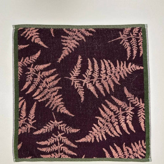 Das Küchenhandtuch "Verzauberter Wald" von Sylvie Thiriez ist ein quadratisches Baumwollhandtuch mit dunklem Hintergrund, rosafarbenem Farnmotiv und dünnem grünen Rand, das flach auf einer hellen Fläche liegt.