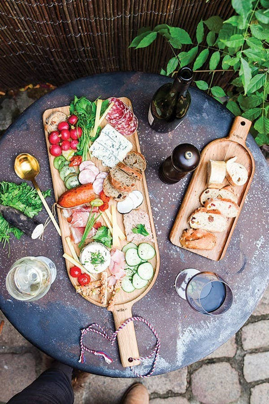 Auf einem runden Tisch im Freien sitzt die LE RÉGAL Große Schaufel, ein Aperitifbrett aus französischem Eichenholz, belegt mit Käse, Fleisch, Brot, Radieschen, Gurken & Grünzeug - ideal für den nachhaltigen Genuss zum Wein.