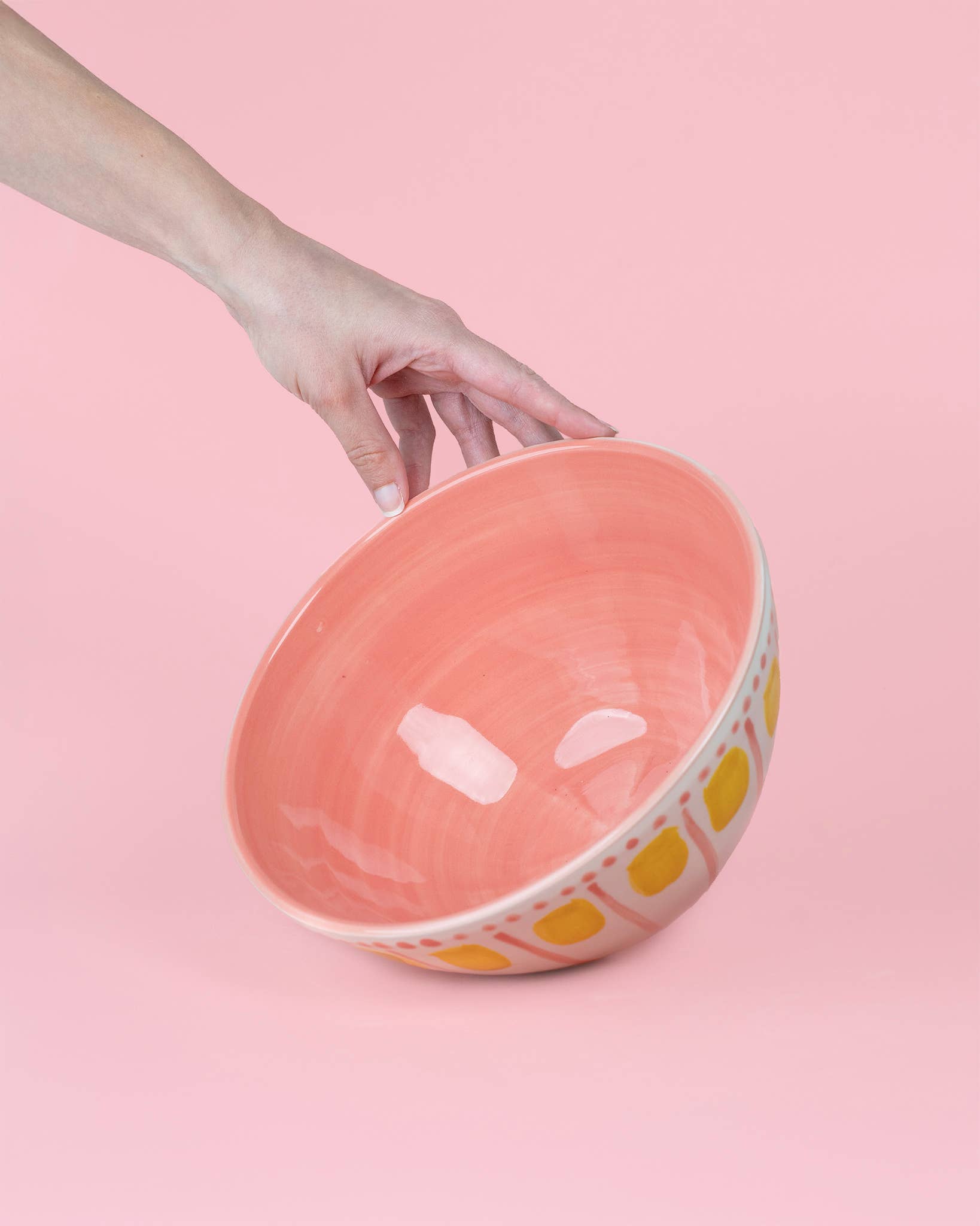 Eine Hand präsentiert die Alfar Tierra Cocida | Handgefertigte Keramik-Servierschale 1,5L VALENTINA in Rosa und Gelb, mit spanischer Handwerkskunst, vor einem rosa Hintergrund.