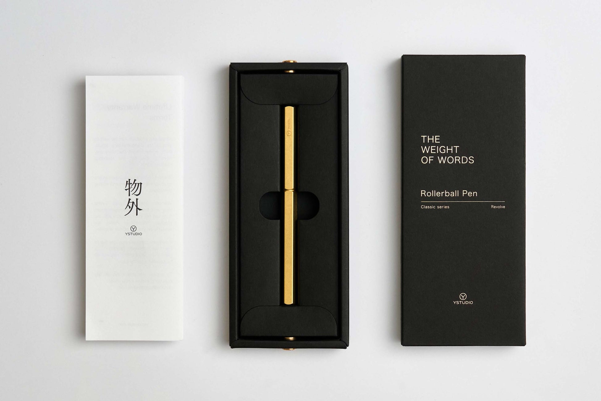 Ein goldener ENTREPÔT - YSTUDIO Klassischer Revolve-Rollerball-Stift (Messing) mit stabilem Griff liegt in einer schwarzen Schachtel zwischen einem weißen japanischen Heft und einem schwarzen Deckel mit der Aufschrift THE WEIGHT OF WORDS. Auf beiden Deckeln ist die Marke ENTREPÔT zu sehen.