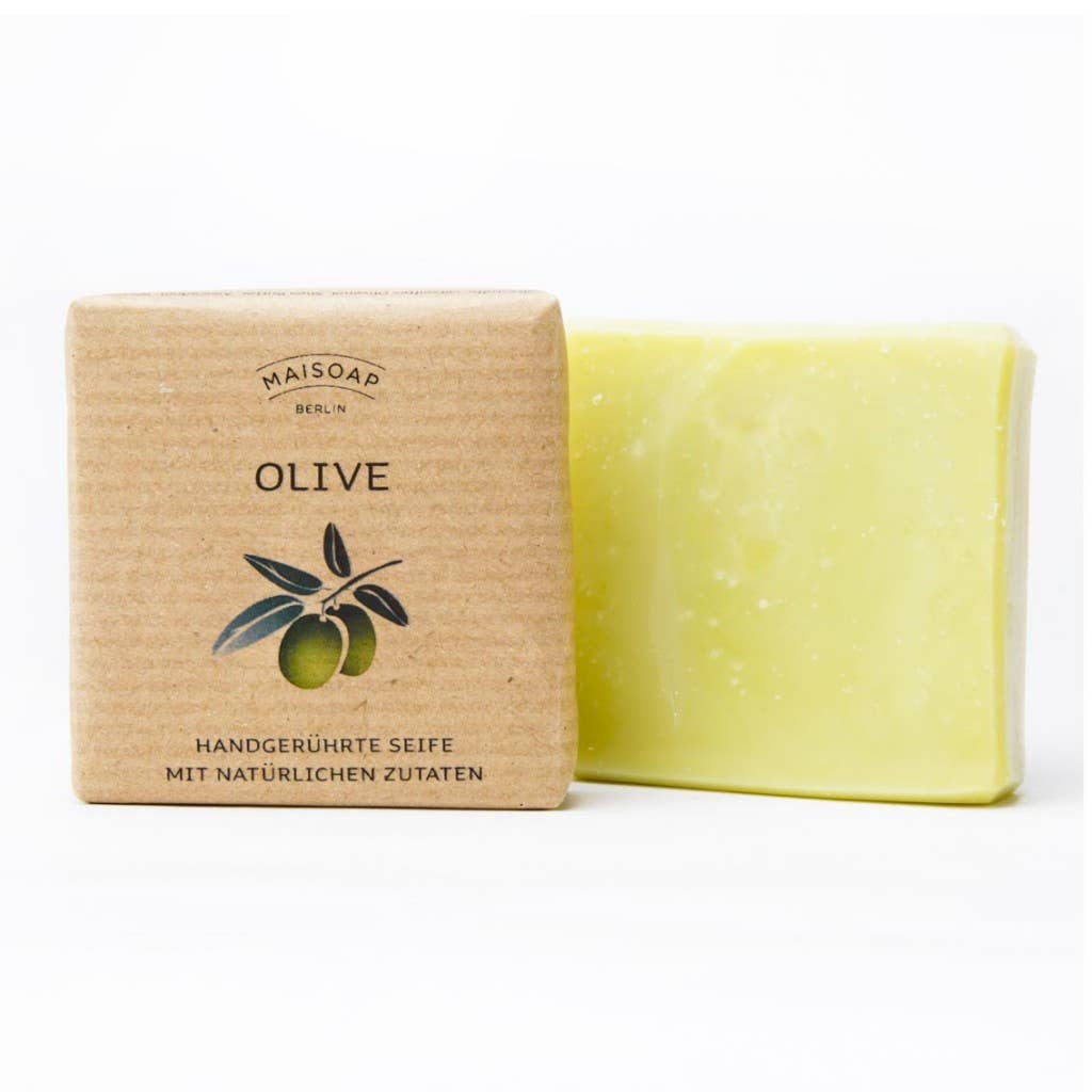 Ein Stück hellgrüne Oliven Seife von maisoap berlin mit Avocadoöl ruht neben der braunen Papierverpackung mit der Aufschrift "Olive", die eine Oliven-Illustration und einen deutschen Text über handgemachte Seife mit natürlichen Inhaltsstoffen zeigt.