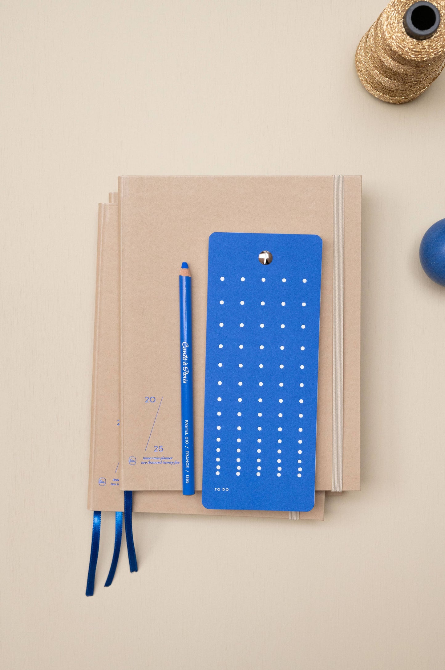 Die Tinne Mia - Aufgabenliste - True Blue von Tinne Mia liegt mit blauem Ribbon-Lesezeichen, passendem Bleistift und perforiertem Lineal auf einer beigen Fläche. Goldene Kordel und blaue Kugel ergänzen das Arrangement.