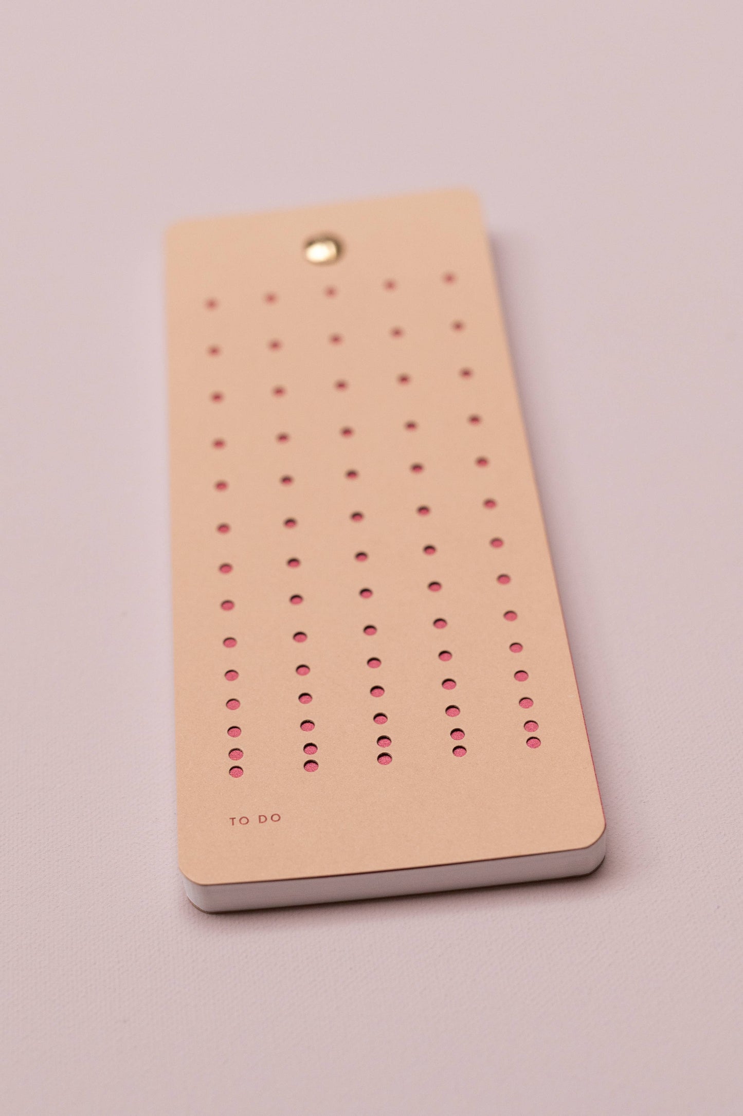 Die Tinne Mia - Aufgabenliste - Nougat von Tinne Mia ist ein rechteckiger beigefarbener Notizblock aus FSC-zertifiziertem Papier mit kleinen rosa Löchern in Reihen, einer Metallniete oben und "TO DO" in der linken unteren Ecke. Abgebildet auf einer hellen Oberfläche.