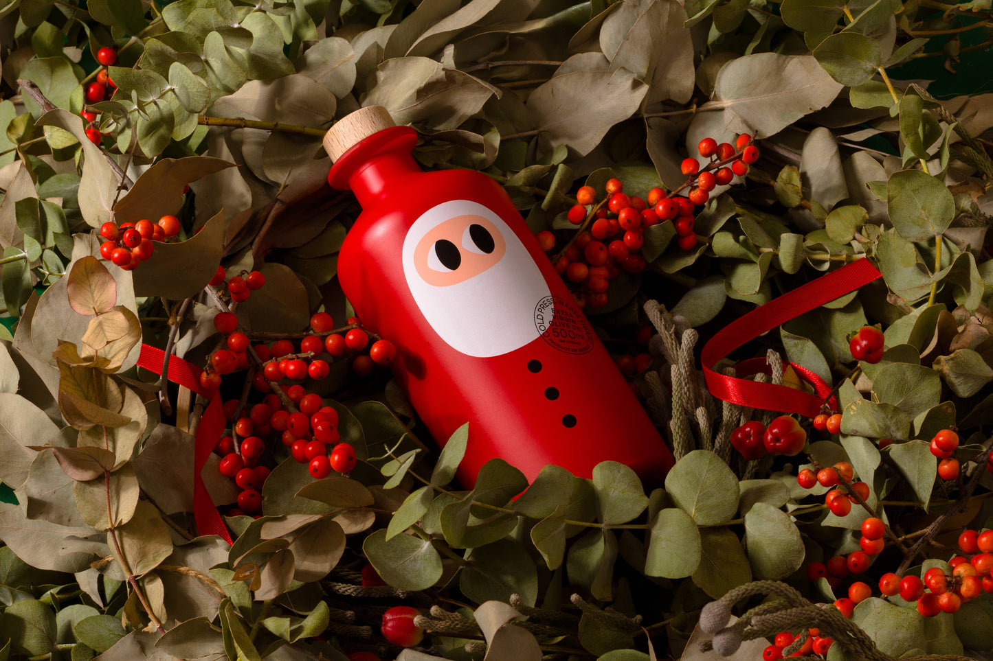 Eine 500 ml Weihnachtsedition von yiayia and friends Natives Olivenöl extra mit Cartoon-Label und Holzverschluss liegt zwischen roten Beeren und Blättern - ein exklusives Geschenk für Feinschmecker und Sammler.