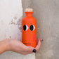 Eine Hand hält eine Orangenflasche mit Augen und einem Korken, perfekt für Yiayia und Freunde - Natives Olivenöl extra mit Chili.