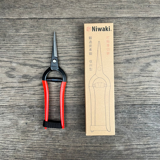 Eine geschmiedete Niwaki-Schere mit roten Griffen liegt neben ihrer braunen Niwaki-Box, die eine Strichzeichnung der Schere sowie englischen und chinesischen Text zeigt, alles angeordnet auf einer Holzoberfläche.