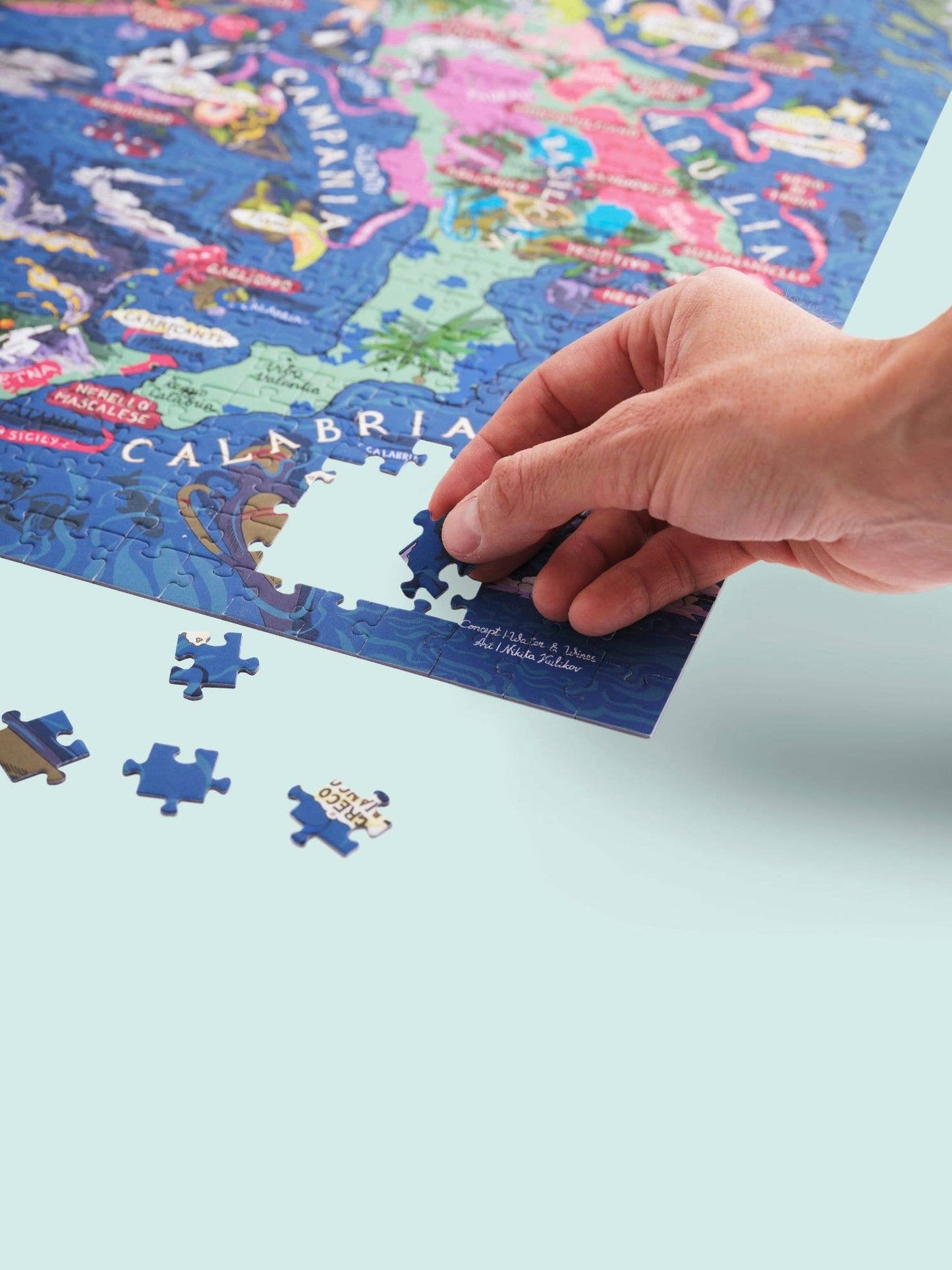 Eine Hand vervollständigt das Water & Wines - Wein Puzzle - Italien, das eine lebendige Karte mit CALABRIA zeigt. Mehrere Teile bleiben auf der hellblauen Oberfläche verstreut und zeigen eine italienische Weinregion.