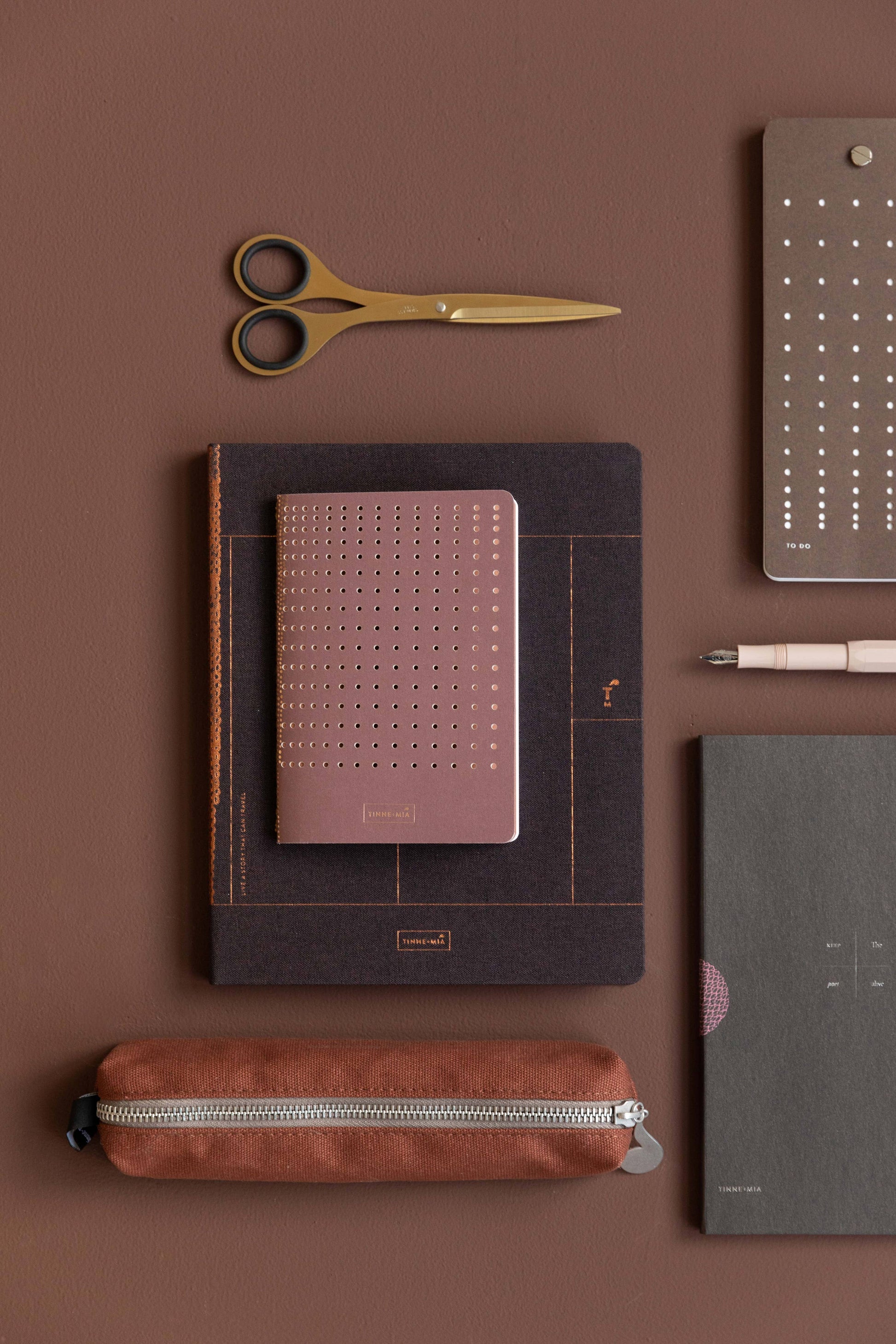 Eine flache Lage brauner und rosafarbener Schreibwaren: eine goldene Schere, ein Stift, ein Notizblock, ein Federmäppchen mit Reißverschluss und das Tinne Mia - Bullet Journal A6 - Grid Burgunderrot - FSC Mix von Tinne Mia, alles fein säuberlich auf einer braunen Fläche angeordnet.