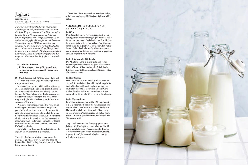 Eine Auswahl von Speisekammer-Glasflaschen und -Gläsern aus dem Hölker Verlag, gefüllt mit Milch und Joghurt, arrangiert auf einer weißen Fläche neben einer aufgeschlagenen Kochbuchseite mit deutschen Joghurt-Rezepten - ideal für Liebhaber selbstgemachter Speisen.