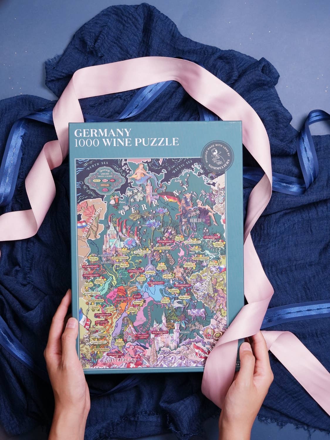 Eine Person hält eine Schachtel Water & Wines - Wein Puzzle - Deutschland (1000 Teile) auf einem dunkelblauen Tuch, mit einem rosa Band um die Schachtel drapiert, vor einem blauen Hintergrund.