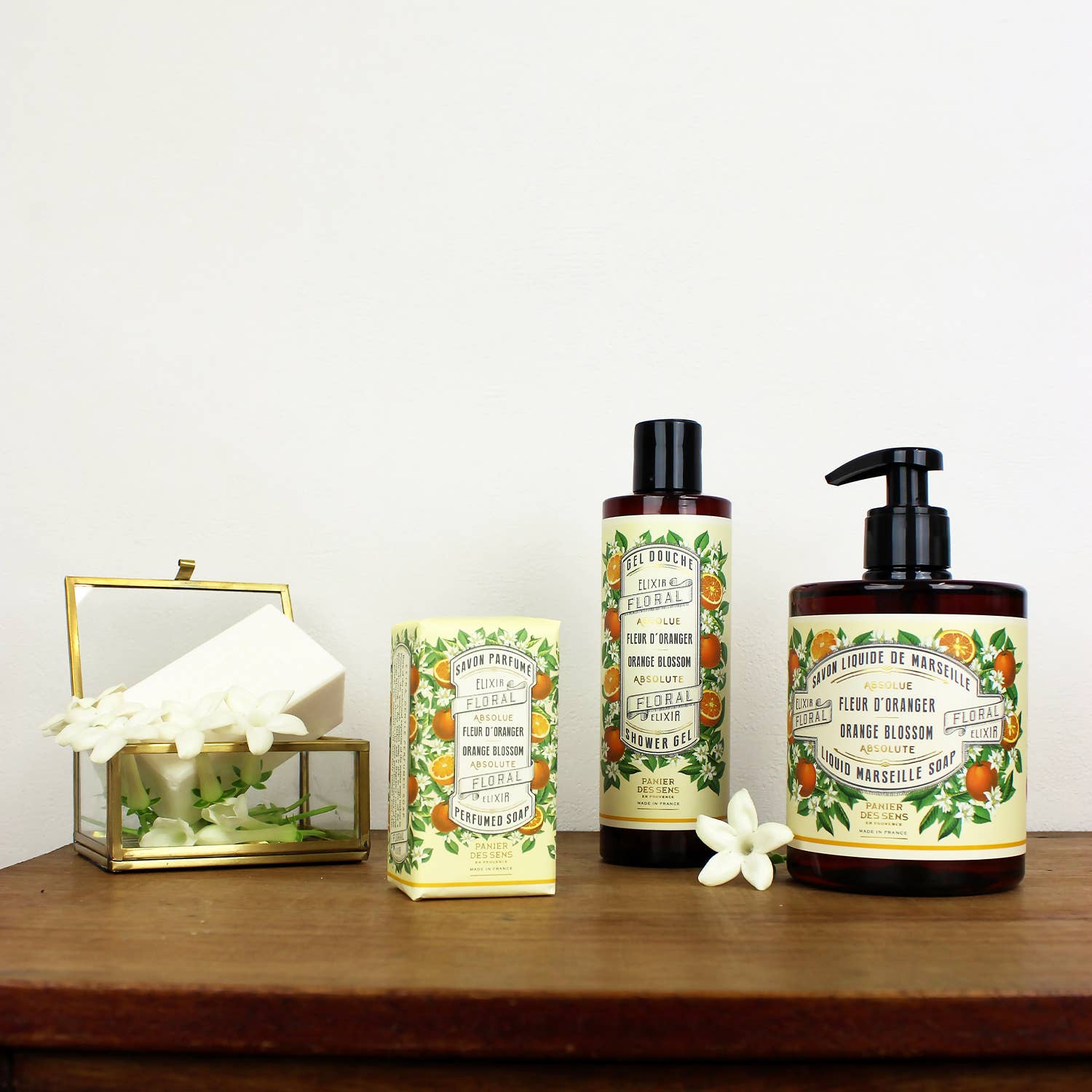 Vier Panier Des Sens Duftseife | Orangenblüte 150 g Seifen mit natürlichen Inhaltsstoffen, in verschiedenen Formen, werden auf einer hölzernen Oberfläche präsentiert, die mit kleinen weißen Blumen verziert ist.