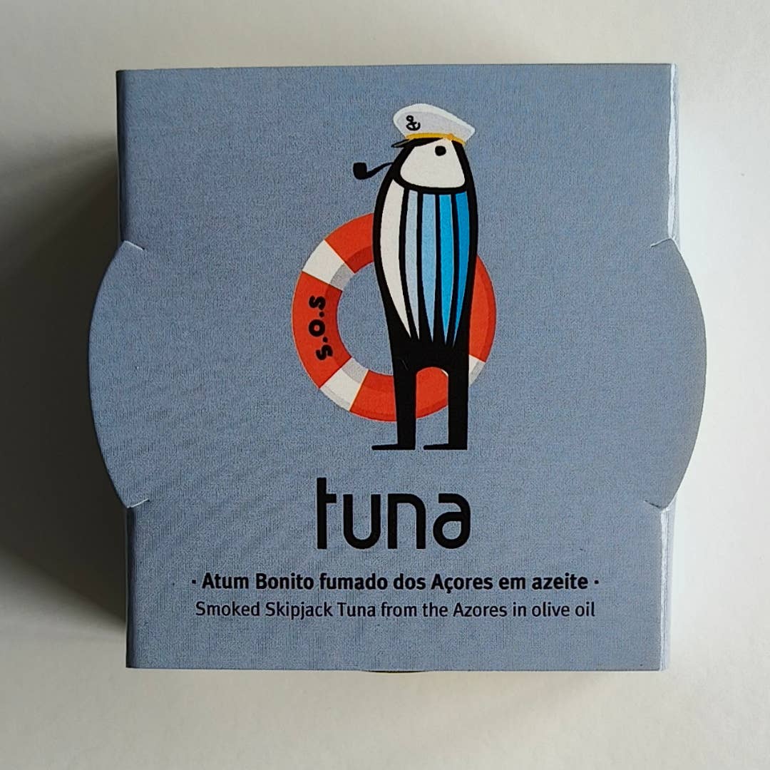 Eine blaue Dose Sardinha - Geräucherter Bonito-Thunfisch aus den Azoren in Olivenöl zeigt einen Cartoon-Thunfisch mit Matrosenhut an einem Rettungsring. Darunter steht "Sardinha - Geräucherter Bonito-Thunfisch aus den Azoren in Olivenöl", dazu Angaben auf Portugiesisch und Englisch.