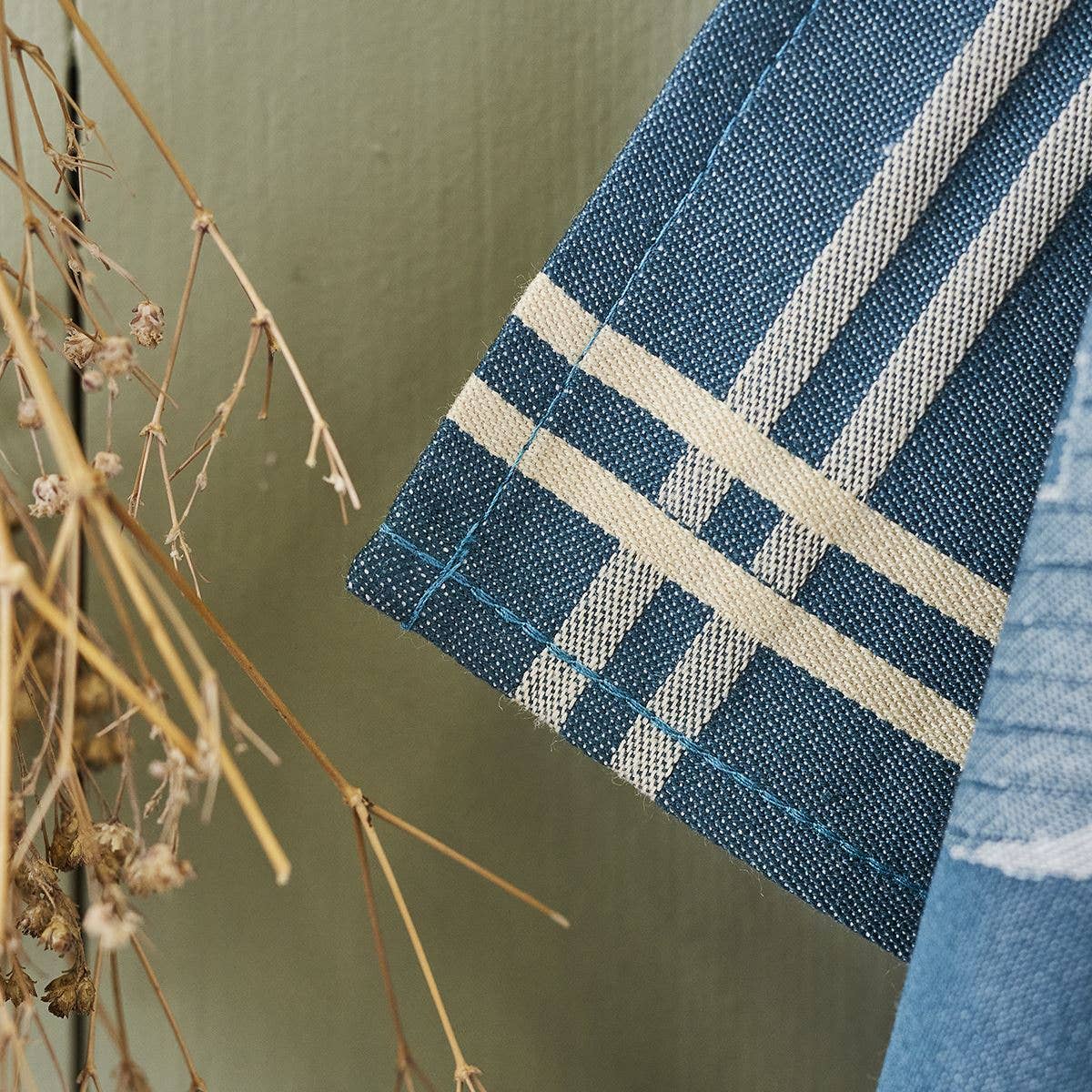 Eine Nahaufnahme der Sylvie Thiriez - Torchon Jacquard Coeur Vallée Blau, OEKO-TEX-zertifizierte Baumwollhandtücher mit weißen Streifen, die neben trockenen beigen Stängeln an einer hellen olivgrünen Wand hängen. Marke: Sylvie Thiriez.