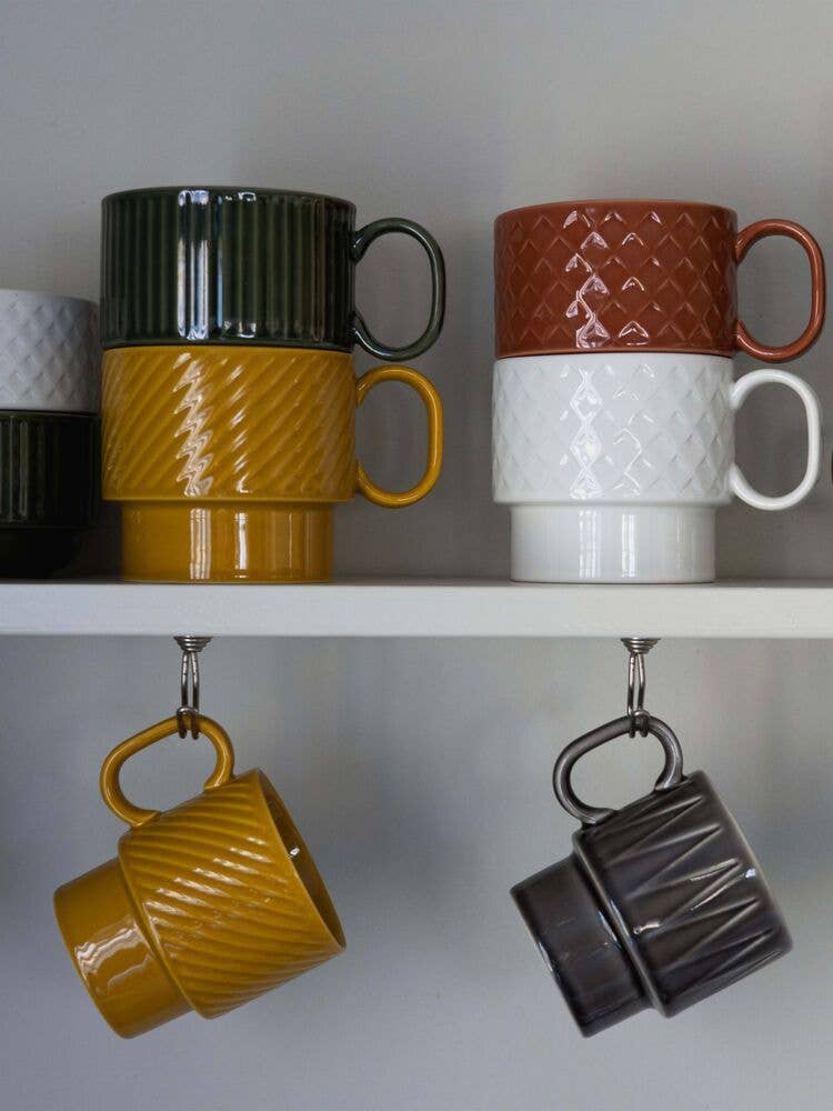 Sechs strukturierte Teetassen der Sagaform GmbH Coffee & More in Grün, Senfgelb, Braun und Weiß sind auf einem weißen Regal gestapelt. Zwei weitere - gelb und dunkelgrau - hängen kopfüber. Genießen Sie besondere Coffee & More Momente mit der Sagaform Coffee & More Teetasse, 2-tlg.