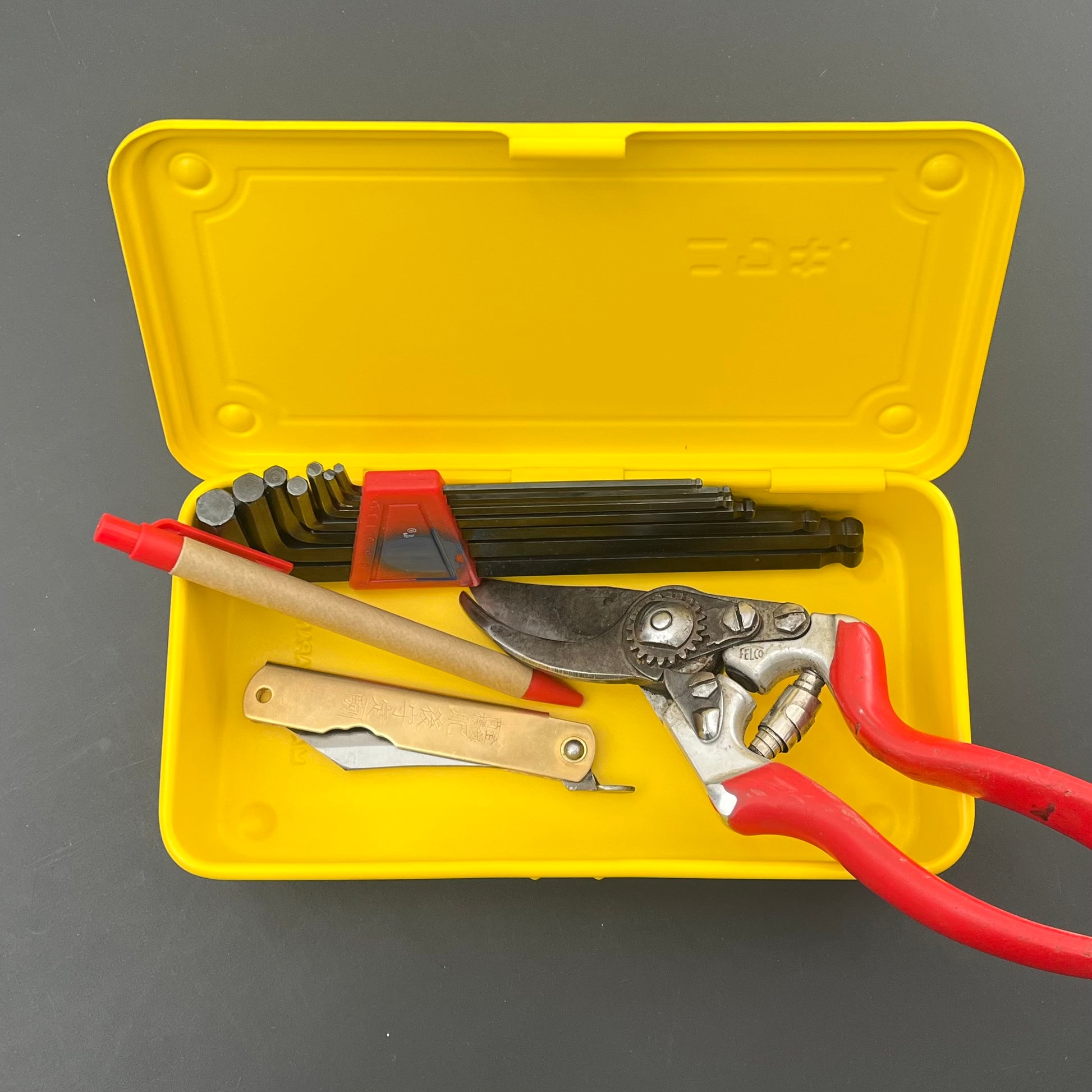 Der gelbe Niwaki T-Type-Werkzeugkasten ist perfekt für Gärtner und Heimwerker und enthält eine Gartenschere mit rotem Griff, ein klappbares Universalmesser, einen roten Marker und Inbusschlüssel.