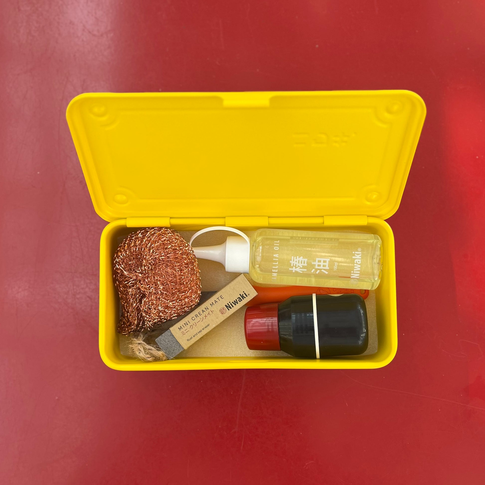 Eine gelbe Schachtel auf einer roten Fläche enthält einen kupfernen Schrubber, ein Stück Seife, den Niwaki Mini Crean Mate, eine klare gelbe Flüssigkeitsflasche mit weißer Pumpe und eine schwarze Flasche mit rotem Verschluss.