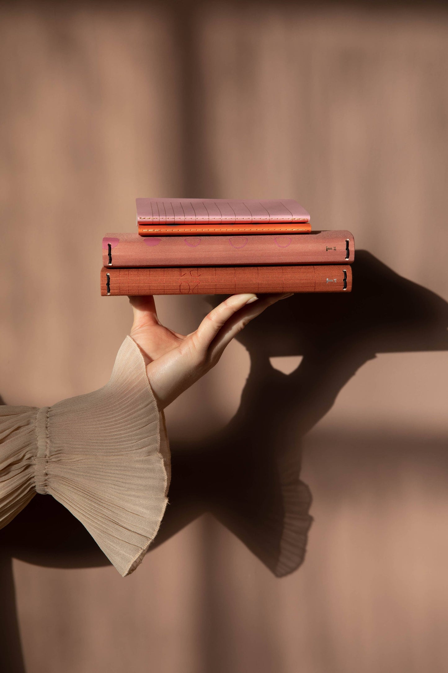 Eine Hand in einem durchsichtigen, plissierten Ärmel hält zwei geschlossene, rotbraune Bücher und das Tinne Mia Notizbuch mit Knopf - Leinen - Rose Dawn FSC Mix, übereinander gestapelt, vor einem sanft beleuchteten, neutralen Hintergrund mit Schattenwurf im Hintergrund.