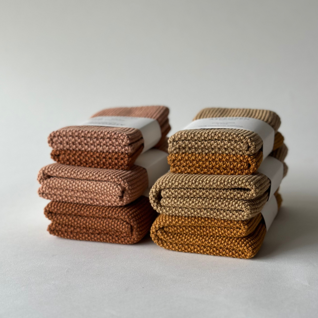 Sechs ordentlich gefaltete Aspegren Denmark - Spültücher, gestrickt, einfarbig, kürbisfarben, liegen in zwei Stapeln auf einer weißen Unterlage, jeweils mit einem weißen Papierband befestigt. Der Hintergrund ist schlicht und hell. Marke: Aspegren Denmark.