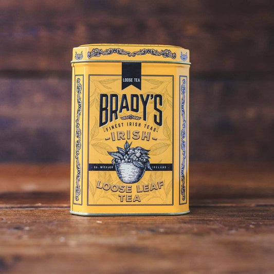 Eine gelbe Dose Brady's Coffee Company – Bradys Loose Leaf Tea, gemischt mit kenianischem Schwarztee vom Kapchorua Estate, zeigt ein kunstvolles Etikett mit eleganten Mustern und der Abbildung einer Teetasse auf einer Holzoberfläche vor einem rustikalen Hintergrund.