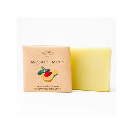 Ein Riegel maisoap berlin - Avocado Minze Seife liegt neben seiner braunen Papierverpackung, die mit Avocado und Minze illustriert ist und auf der auf Deutsch steht, dass sie mit Avocadoöl handgemacht ist und eine minzige Duftmischung aufweist.