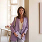 Eine Frau in einem hellvioletten Blazer und weißem Oberteil steht an einer Wand in einem hellen Büro und hält die Tinne Mia Aufgabenliste - Lavendel von Tinne Mia und eine Umhängetasche in der Hand. Im Hintergrund sind ein Schreibtisch und Regale zu sehen.