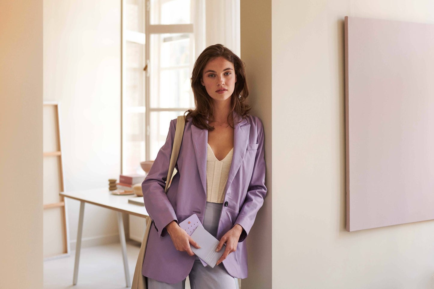 Eine Frau in einem hellvioletten Blazer und weißem Oberteil steht an einer Wand in einem hellen Büro und hält die Tinne Mia Aufgabenliste - Lavendel von Tinne Mia und eine Umhängetasche in der Hand. Im Hintergrund sind ein Schreibtisch und Regale zu sehen.