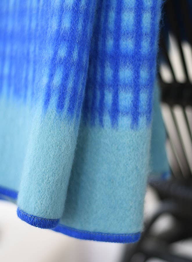 Eine Nahaufnahme der Darling Tagesdecke aus Lammwolle in Blau von Sofie Sjöström Design zeigt das blaue Karomuster, den hellblauen Farbverlauf und den blauen abgesteppten Rand. Der Hintergrund ist unscharf und hebt die weiche Lammwolltextur hervor.