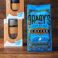 Eine leuchtend blaue Tüte Brady's Coffee Company - Brady's Signature Blend Kaffee liegt auf einer hölzernen Unterlage, während zwei verpackte wiederverwendbare KeepCup-Kaffeebecher im Hintergrund verschwimmen.