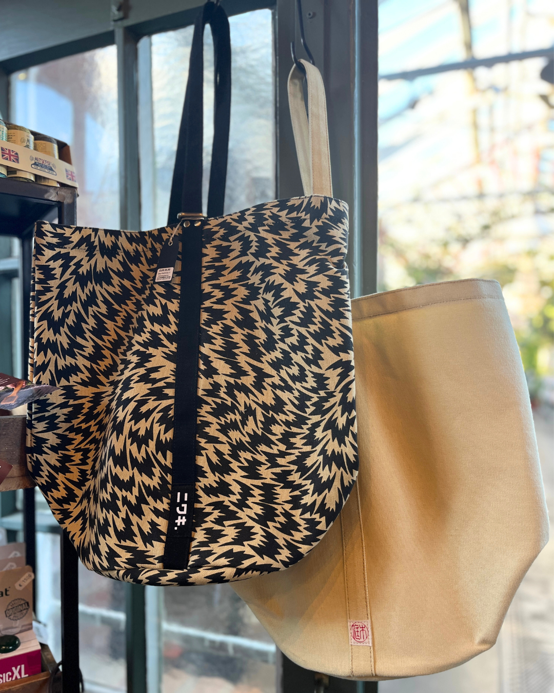 Zwei Tragetaschen hängen an ihren Henkeln: die Kantan Werkzeugtasche - Niwaki der Königlichen Gartenakademie mit einem auffälligen schwarz-hellbraunen Zickzack-Muster und einem schwarzen Riemen sowie eine schlichte beigefarbene Leinentasche mit einem kleinen roten Logo. Dahinter sind Fächer und ein Fenster zu sehen.