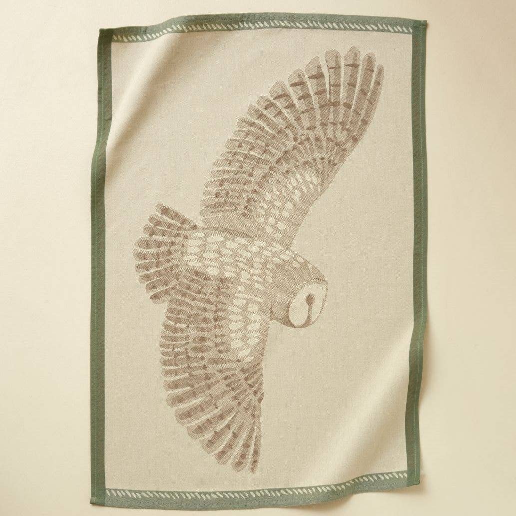 Das Sylvie Thiriez - Geschirrtücher Jacquard L'envol naturel von Sylvie Thiriez ist ein beigefarbenes Baumwoll-Küchentuch mit grünem Rand, auf dem eine Schleiereule im Flug abgebildet ist.