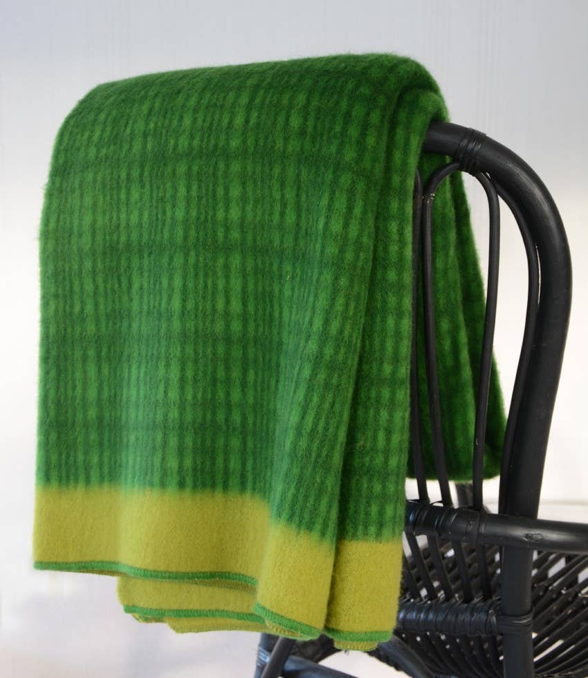 Die Darling Tagesdecke aus Lammwolle | Kiwi von Sofie Sjöström Design, eine grün karierte Wolldecke mit gelber Umrandung, ist über die Rückenlehne eines schwarzen geflochtenen Stuhls vor einem schlichten hellen Hintergrund drapiert.