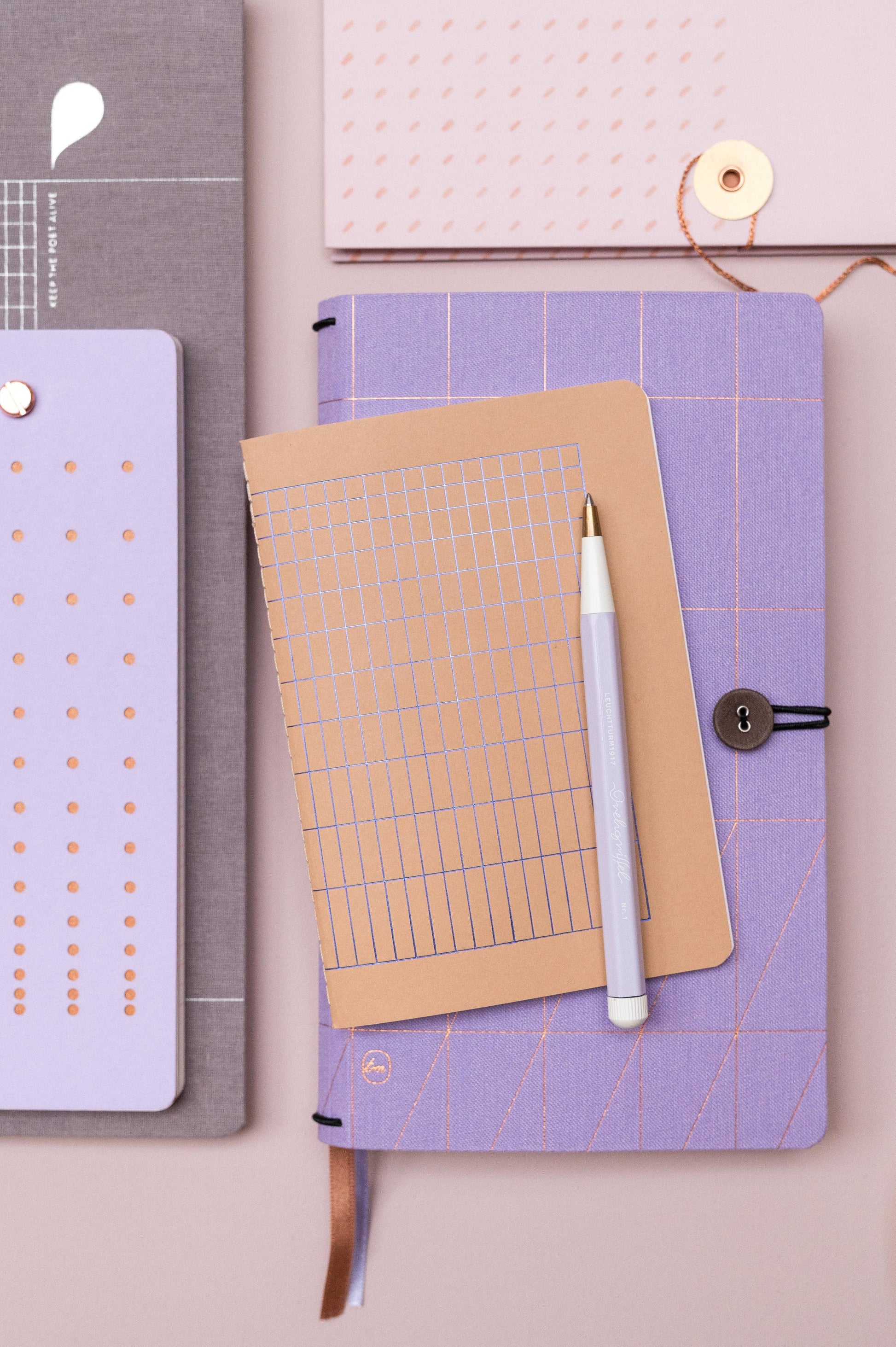 Ein Tinne Mia Notizbuch A6 in Nougat Grid (FSC Mix), ein pastelllila Notizbuch und ein weißer Stift liegen ordentlich auf rosa Unterlage; daneben weitere Hefte mit gepunktetem Raster.