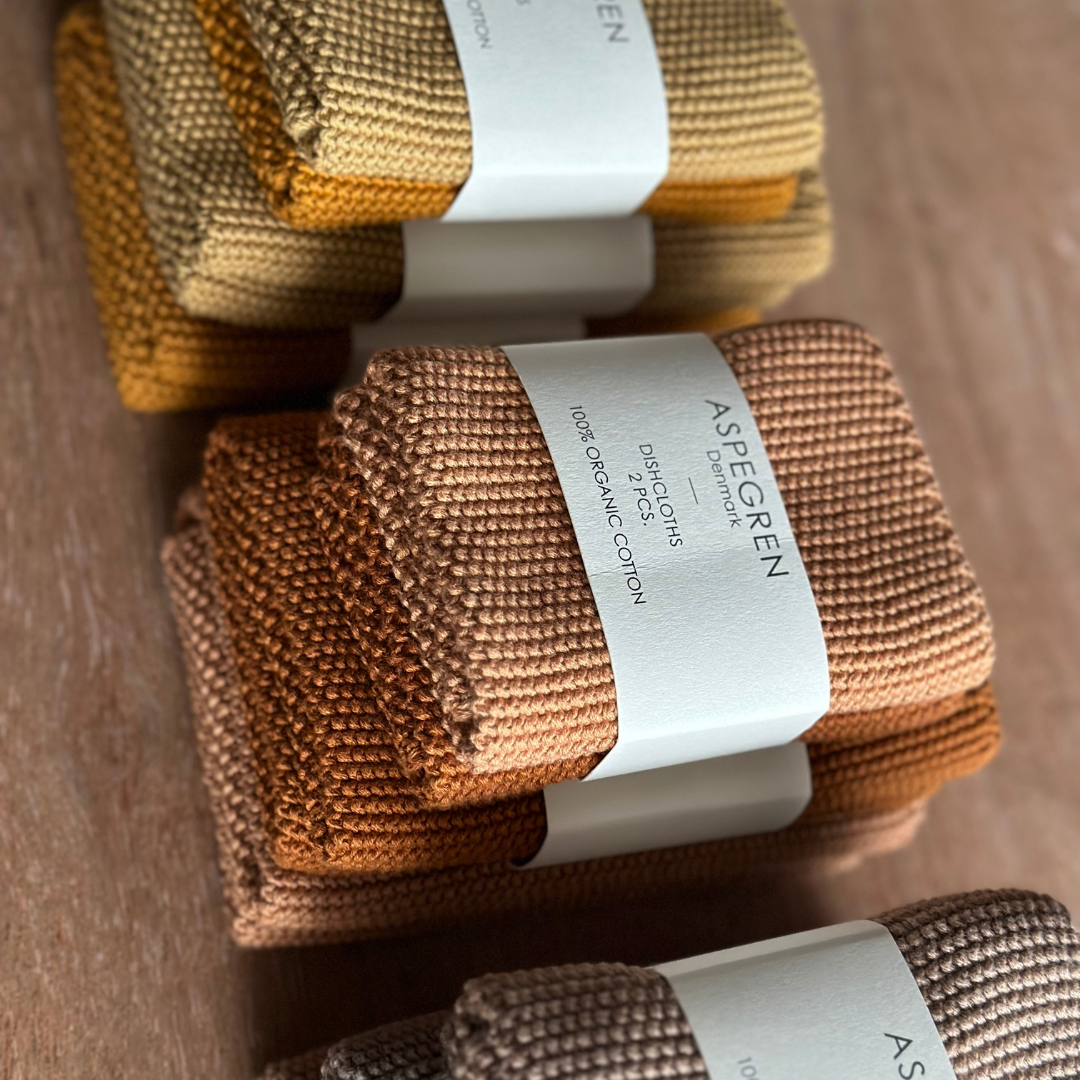 Drei Stapel Spültücher von Aspegren Denmark, gestrickt, einfarbig, kürbisfarben, jeweils mit einem weißen Etikett aus 100 % Bio-Baumwolle von Aspegren Denmark umwickelt, sind auf einer Holzoberfläche angeordnet.