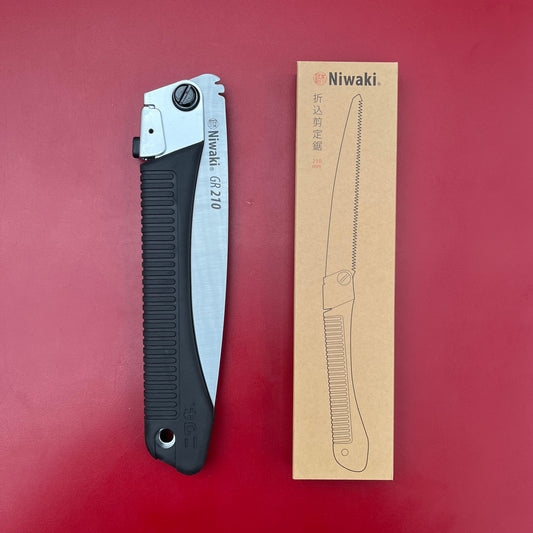 Eine zusammengeklappte Niwaki GR 210 Klappsäge von Niwaki mit schwarzem Griff liegt neben ihrer beigen Schachtel, auf der eine Strichzeichnung der Säge vor rotem Hintergrund zu sehen ist – perfekt für Gartenarbeit und Aufgaben im Freien.